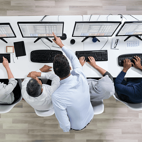 Man pointing to one of his workers screens discussing operating feasibility whilst colleagues next to them continue to type at their computers